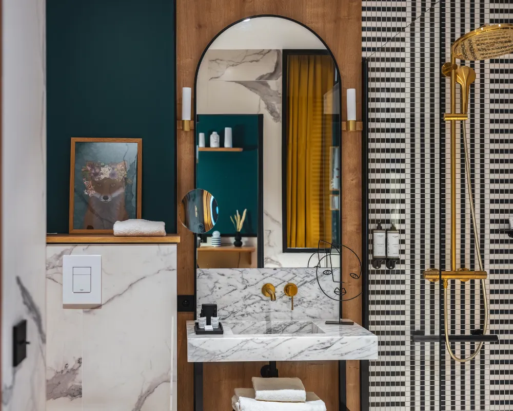 Salle de bain en marbre et bois avec miroir arqué, tableau encadré, vasque à robinetterie dorée et douche vitrée à mosaïque noire et blanche