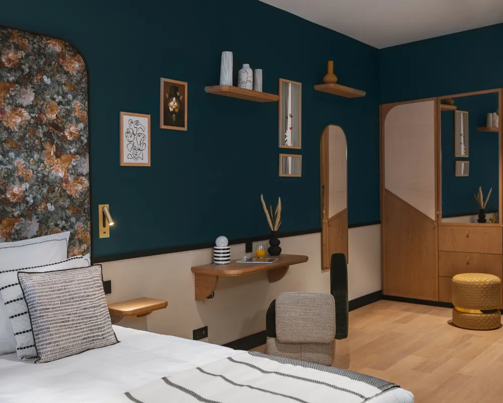 Chambre bleu paon avec tête de lit florale, étagères murales et coin coiffeuse en bois près d'un pouf doré