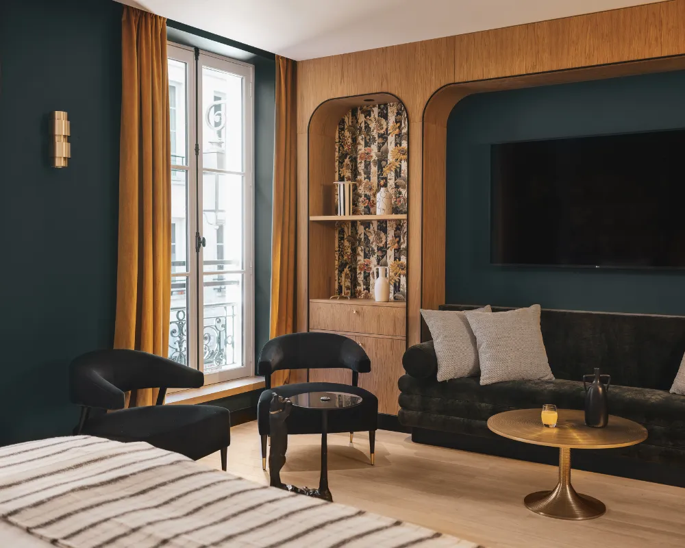 Salon bleu paon avec canapé noir et fauteuils, niche en bois à étagères, télévision murale et fenêtre à balcon parisien