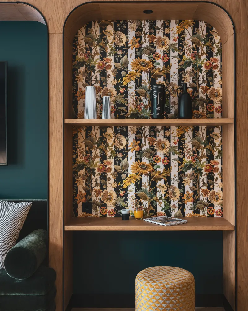 Bibliothèque arquée en bois tapissée de papier peint floral avec bibelots, pouf doré posé en dessous sur fond bleu paon