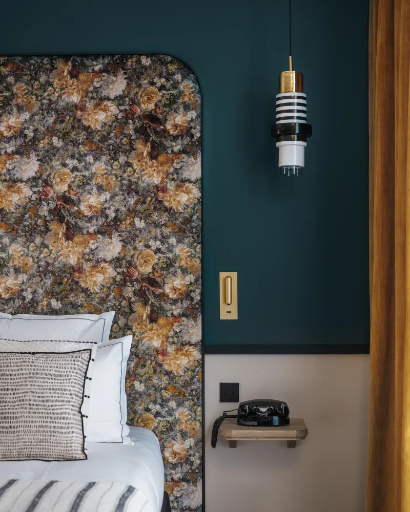 Panneau de tête de lit à motif floral sombre bordé de bleu paon, suspension dorée et téléphone vintage posé sur une tablette murale
