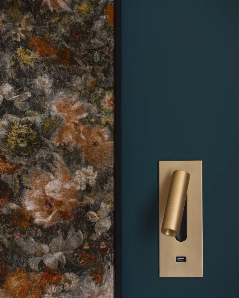 Bordure de panneau floral sombre juxtaposée à un mur bleu paon, liseuse dorée orientable fixée à côté