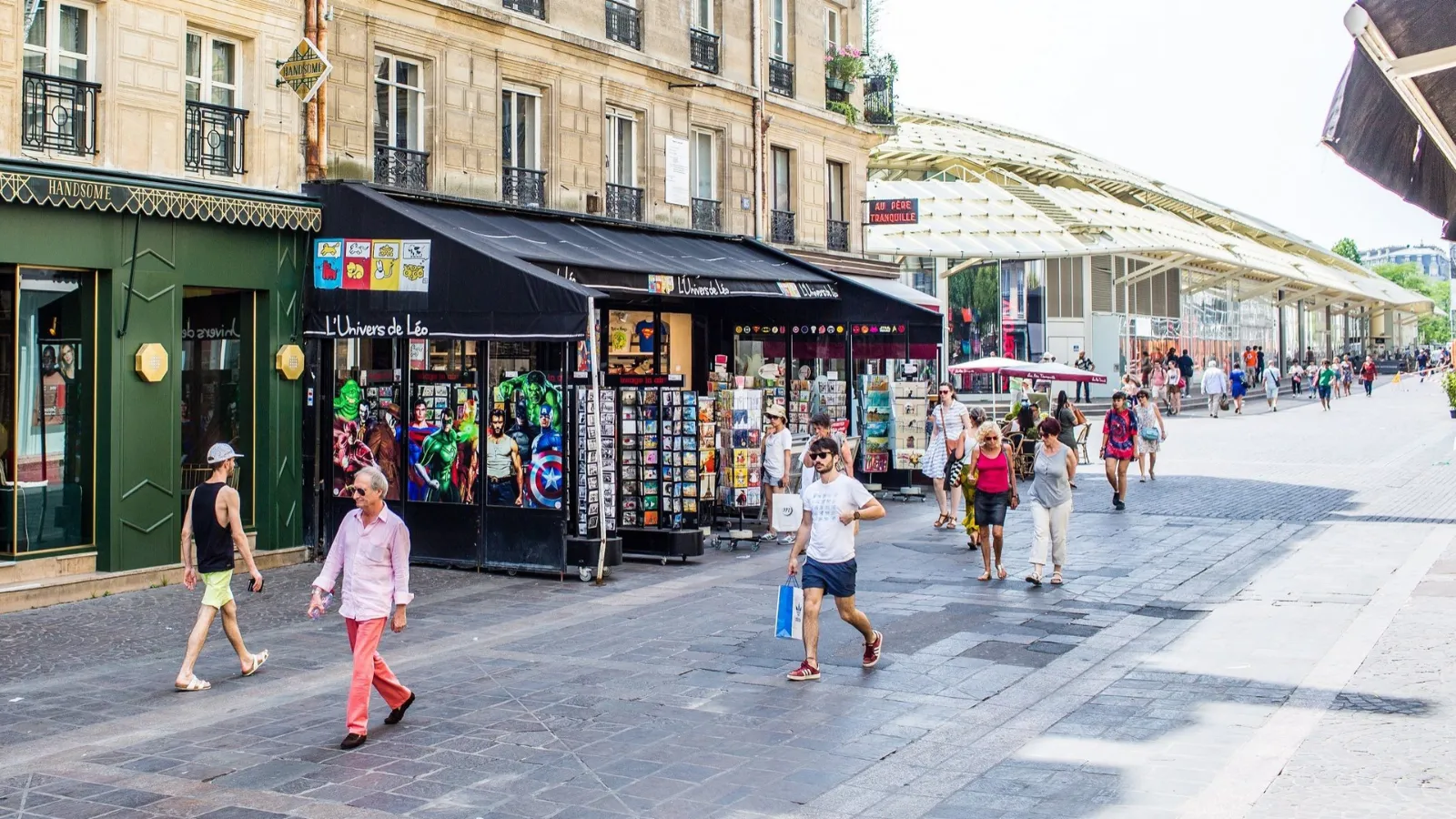 rue pavée ensoleillée avec kiosque et passants, verrière du marché des Halles à l'horizon