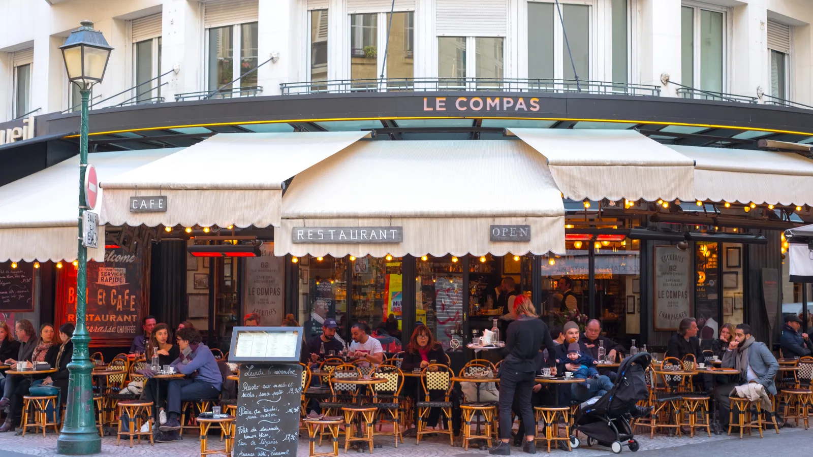 terrasse animée du café Le Compas sous ses auvents crème, clients attablés et réverbère vert au premier plan
