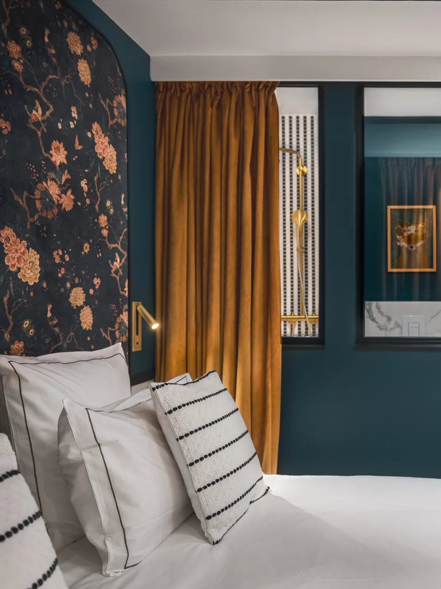Tête de lit florale sombre bordée de bleu paon, rideau moutarde et verrière vitrée sur une douche à robinetterie dorée