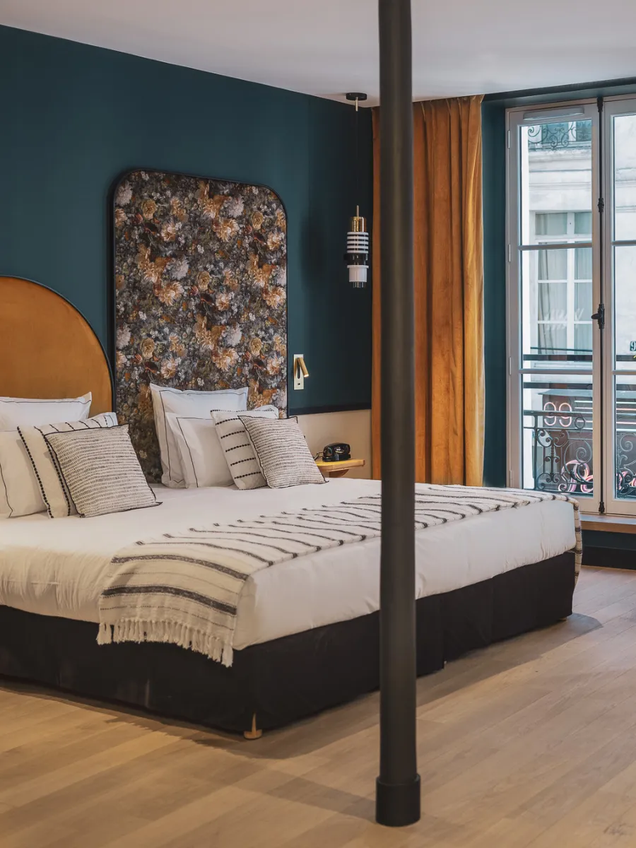Chambre spacieuse au mur bleu paon avec tête de lit florale, colonne de lit à baldaquin et fenêtre ouverte sur un balcon parisien