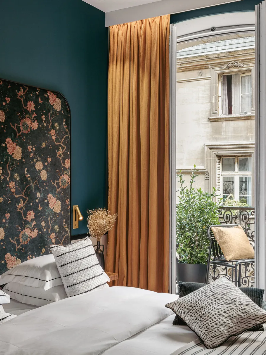 Lit à tête de lit florale sombre bordée de bleu paon, rideau moutarde ouvert sur un balcon en fer forgé face à des immeubles haussmanniens