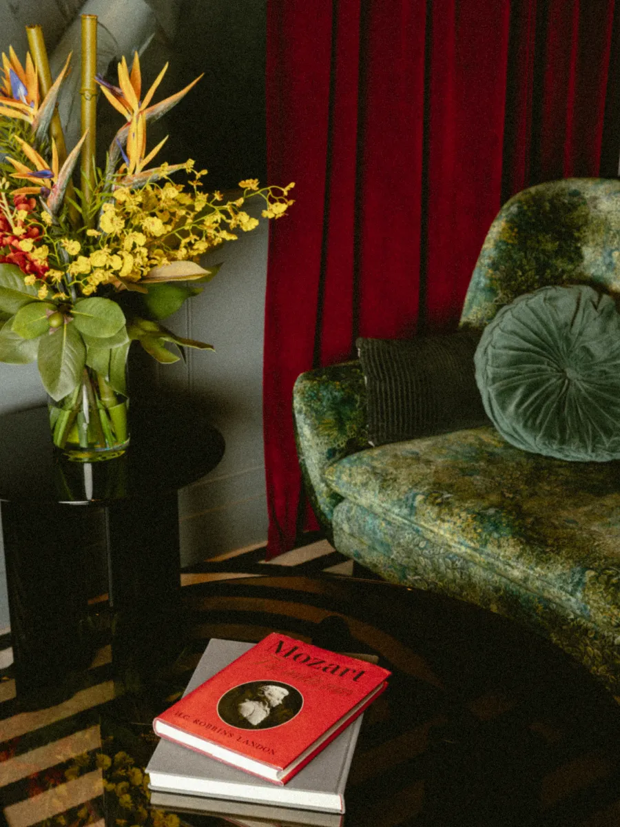 Fauteuil en velours à motifs vert et doré, coussins ronds près d'un bouquet d'oiseaux de paradis et de livres sur Mozart, rideau bordeaux en fond