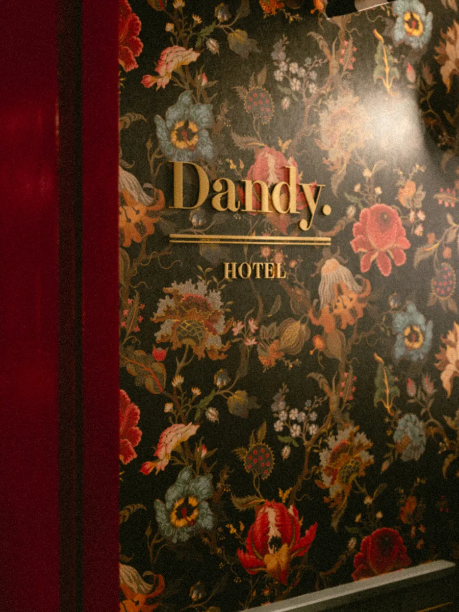 Enseigne dorée Dandy Hôtel fixée sur un papier peint floral sombre, bordée d'un panneau bordeaux laqué