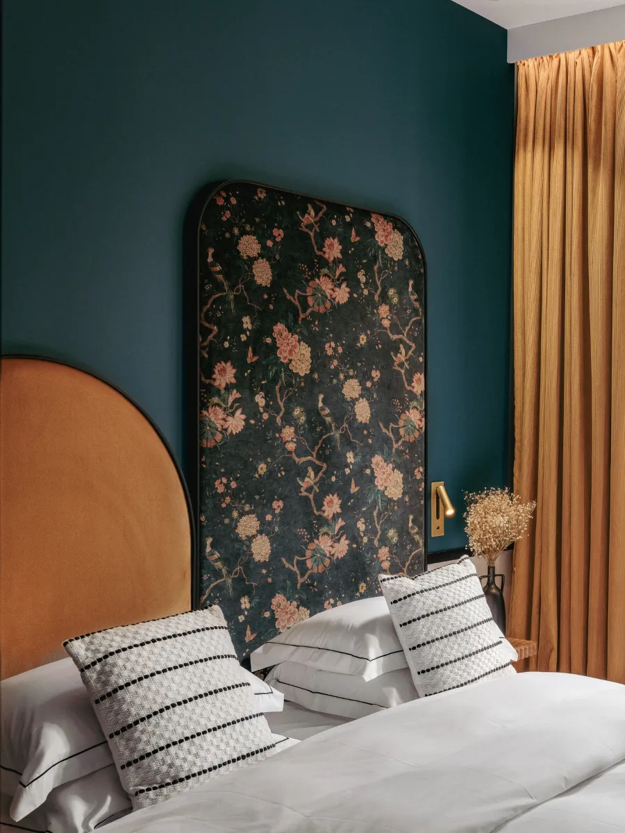 Vue frontale du lit à tête arquée camel et panneau floral bleu nuit, liseuse dorée et bouquet de fleurs séchées devant les rideaux moutarde