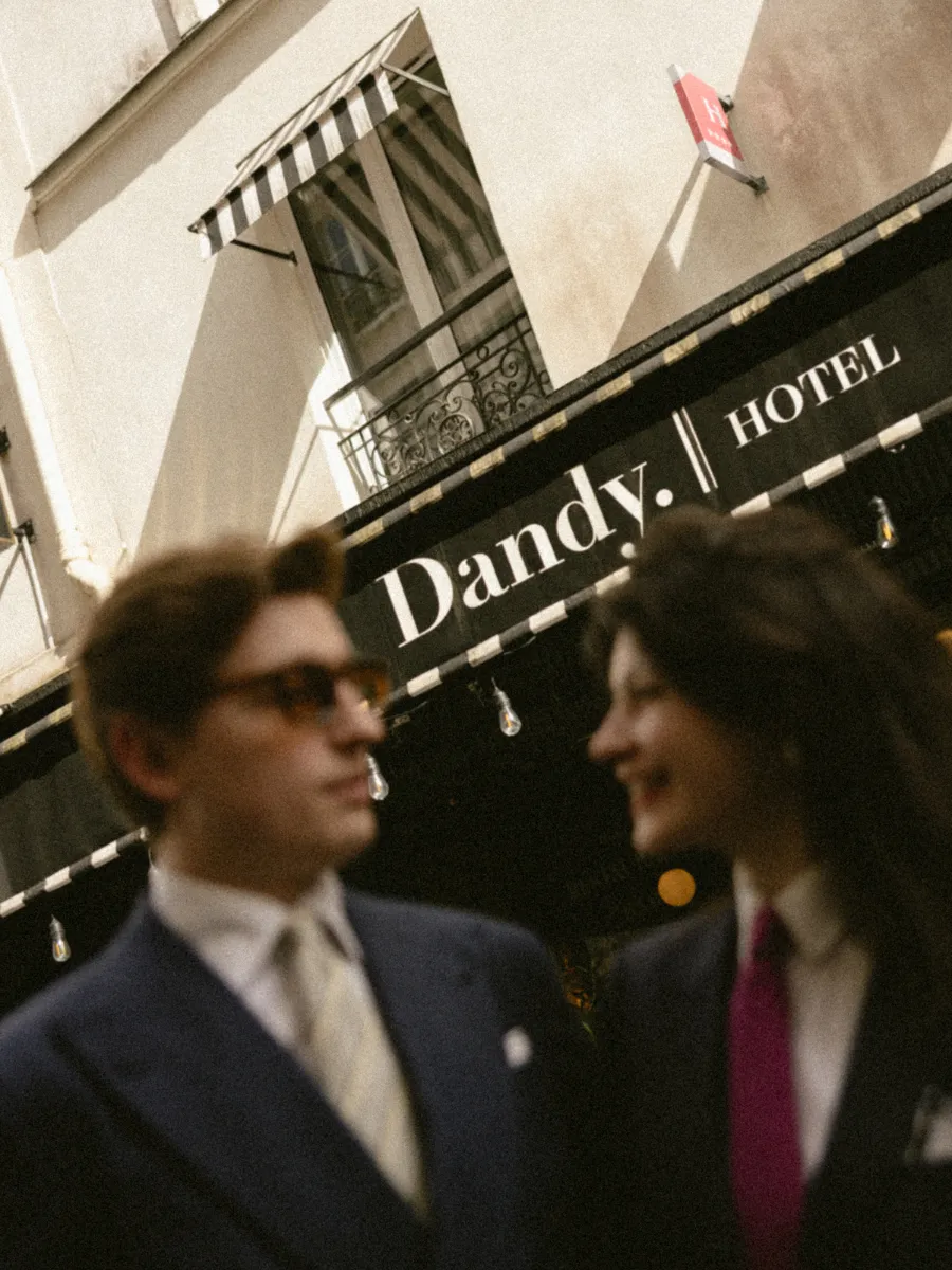 Couple souriant en costume face à face devant l'enseigne rayée du Dandy Hôtel, guirlande d'ampoules suspendue à l'auvent