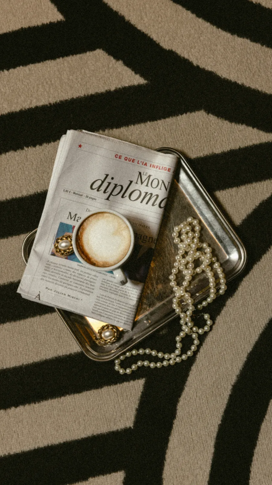 plateau argenté avec tasse de café et journal, collier posé dessus sur le tapis à motifs arqués noir et beige