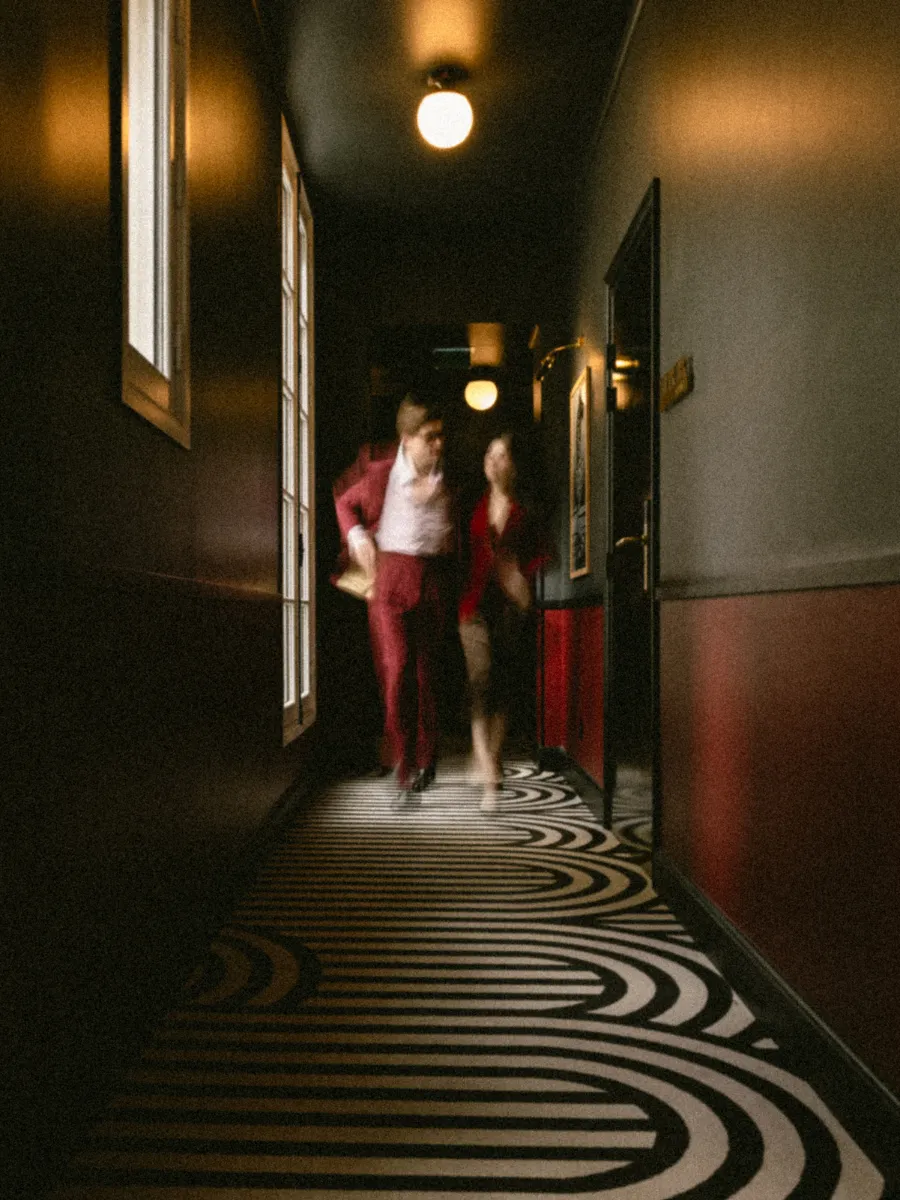 Couple en tenue rouge marchant en flou de mouvement dans le couloir tapissé de motifs arqués, ampoules nues éclairant le passage