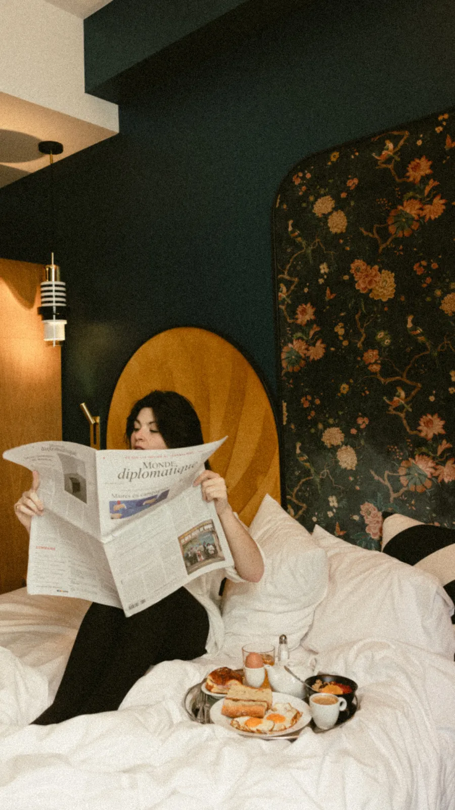 couple prenant son petit-déjeuner au lit, elle lisant le journal sous la tête de lit florale et lui buvant son café