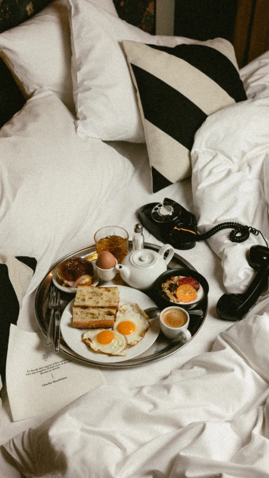 Miniature du plateau argenté de petit-déjeuner sur le lit, coussin rayé noir et blanc et téléphone vintage décroché