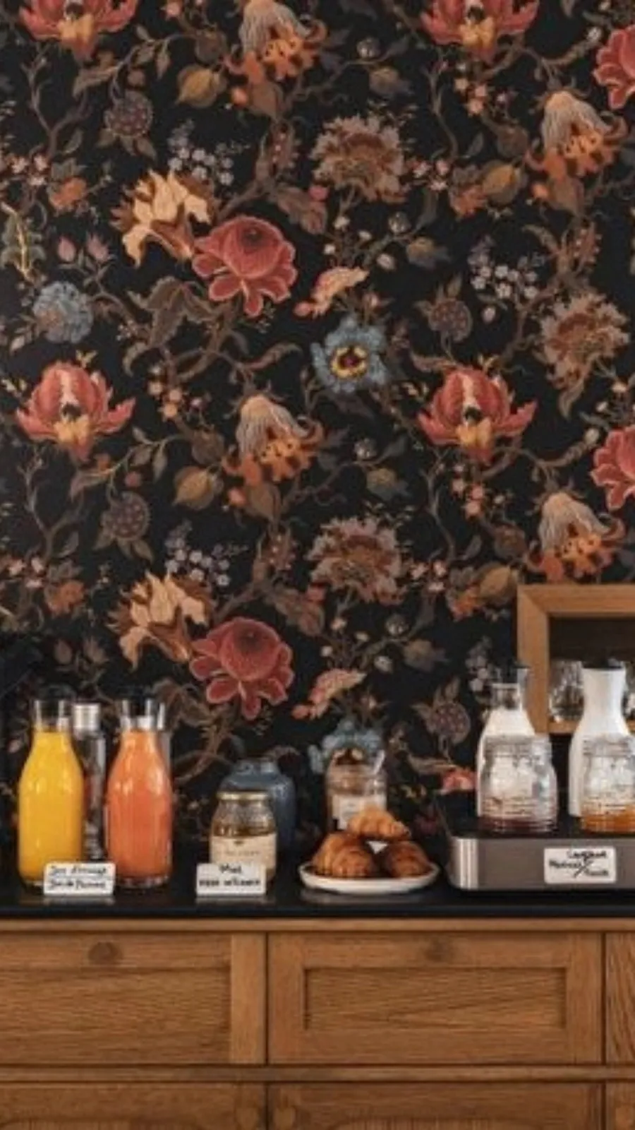 un buffet en bois garni de bouteilles de jus et de céréales, devant un papier peint floral sombre à fleurs orangées