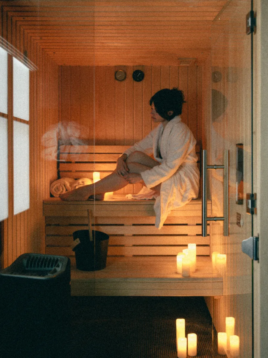 Vue générale du sauna en bois éclairé aux bougies, femme en peignoir assise près des serviettes roulées, photographiée à travers deux vitres