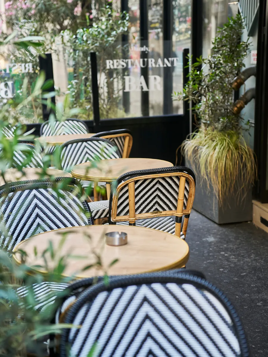 Rangée de chaises bistrot à motifs chevrons devant la vitrine miroir du restaurant bar, cendrier posé sur une table ronde