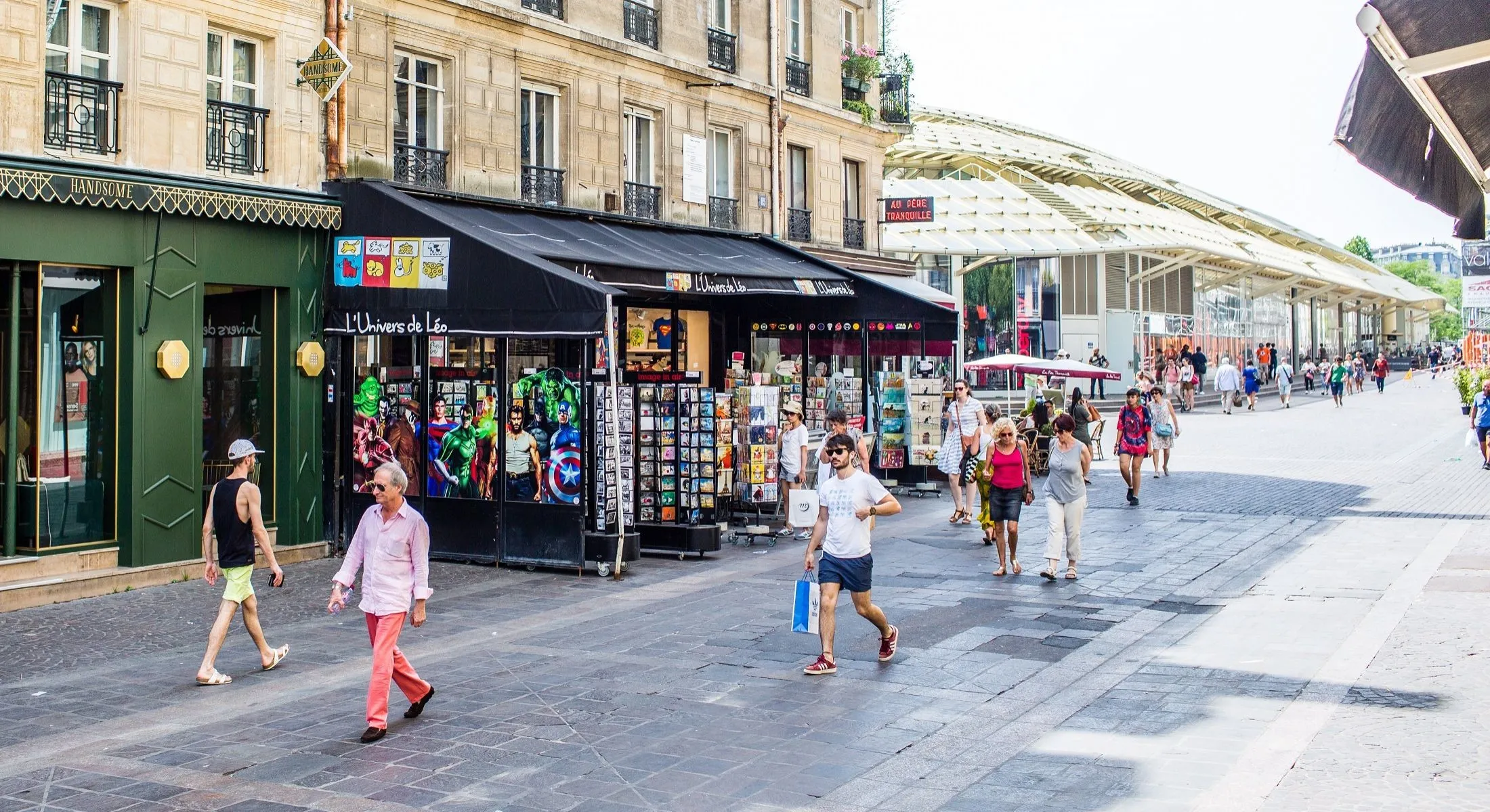 Rue piétonne animée bordée d'échoppes à cartes postales et de boutiques, verrière métallique du marché en arrière-plan