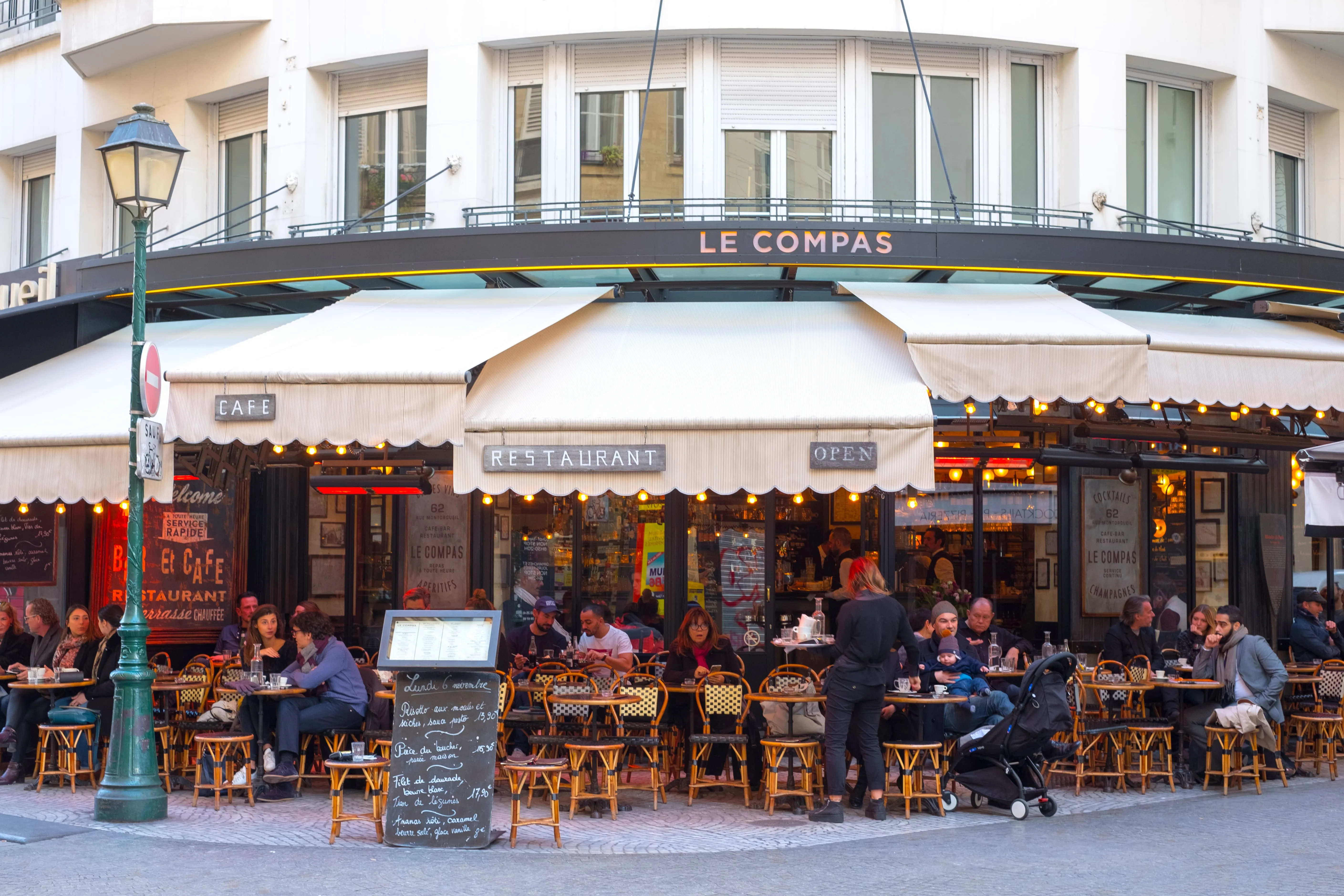 Terrasse bondée du café Le Compas à l'angle d'une rue, serveuse circulant entre les tables sous les auvents beiges