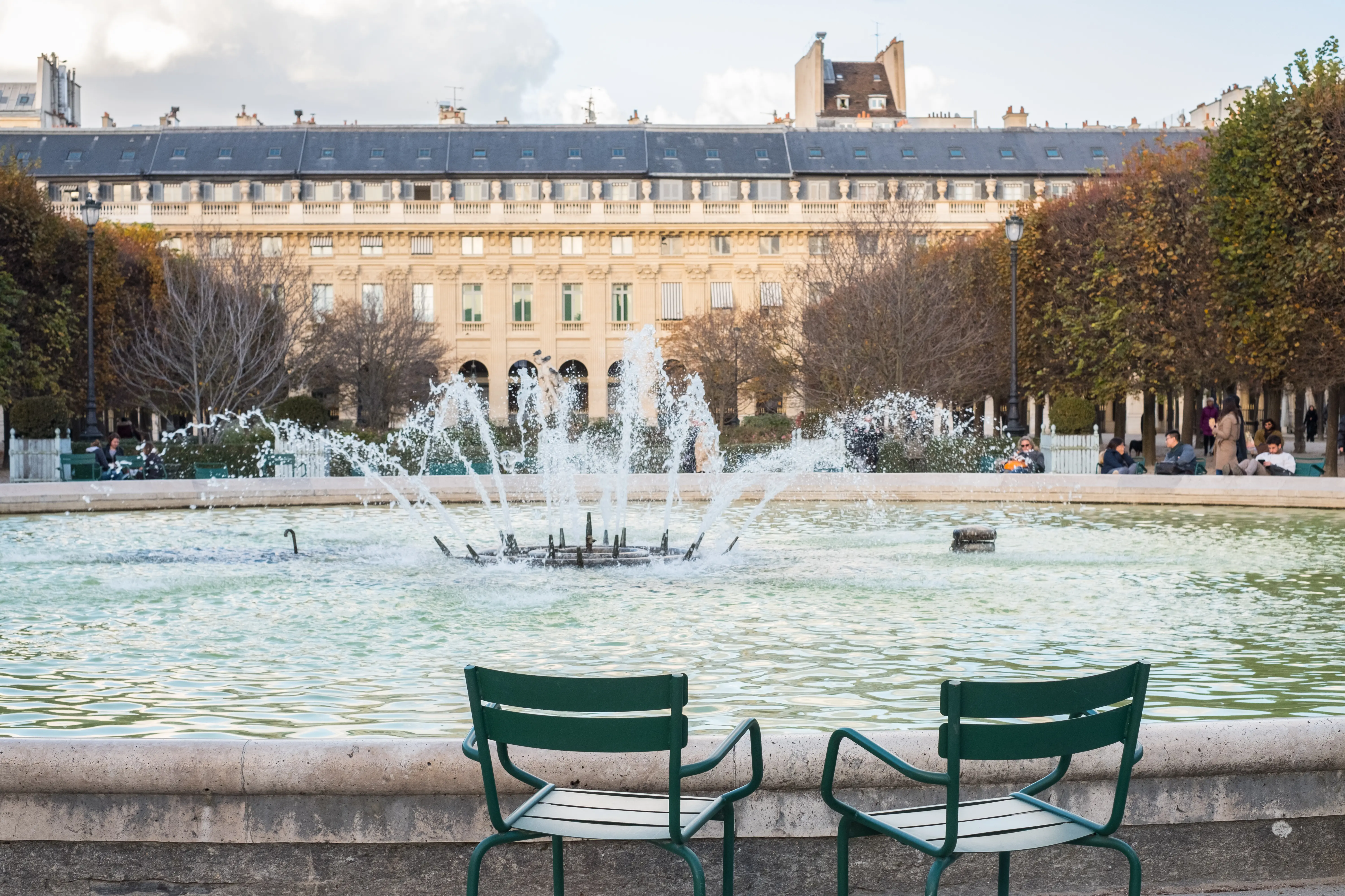 Deux chaises vertes vides face au bassin jaillissant du jardin du Palais-Royal, façade classique en arrière-plan