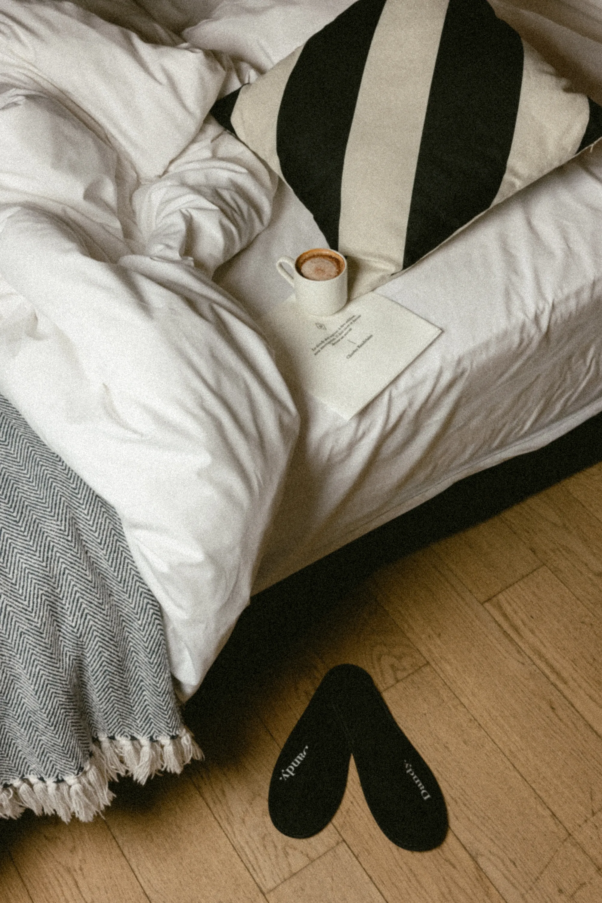 Tasse de café posée sur un lit défait à coussin rayé noir et blanc, chaussons noirs marqués Dandy sur le parquet