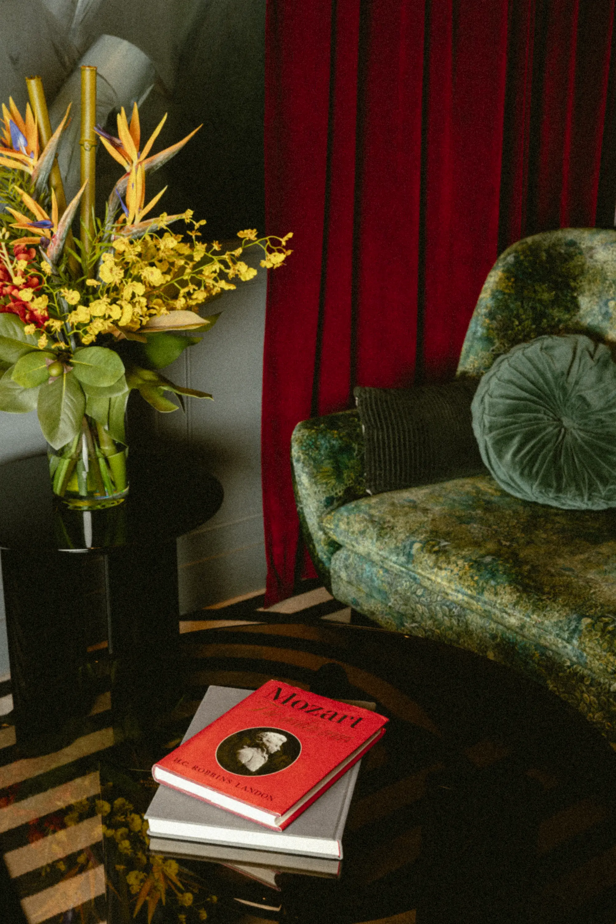 un fauteuil en velours vert à côté d'un bouquet d'oiseaux de paradis, pile de livres posée sur une table basse devant un rideau bordeaux