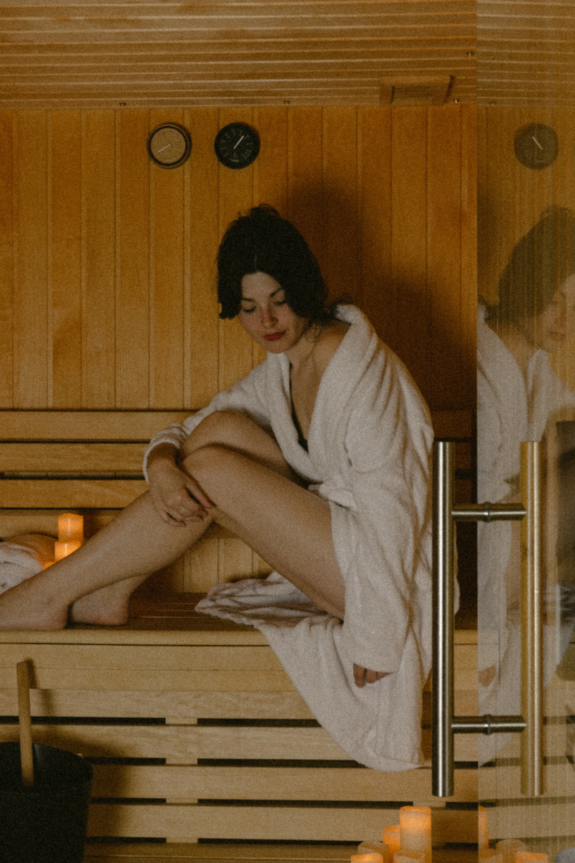 Femme au regard baissé assise dans le sauna en bois, chignon défait et peignoir glissant sur l'épaule, reflet dédoublé dans la vitre