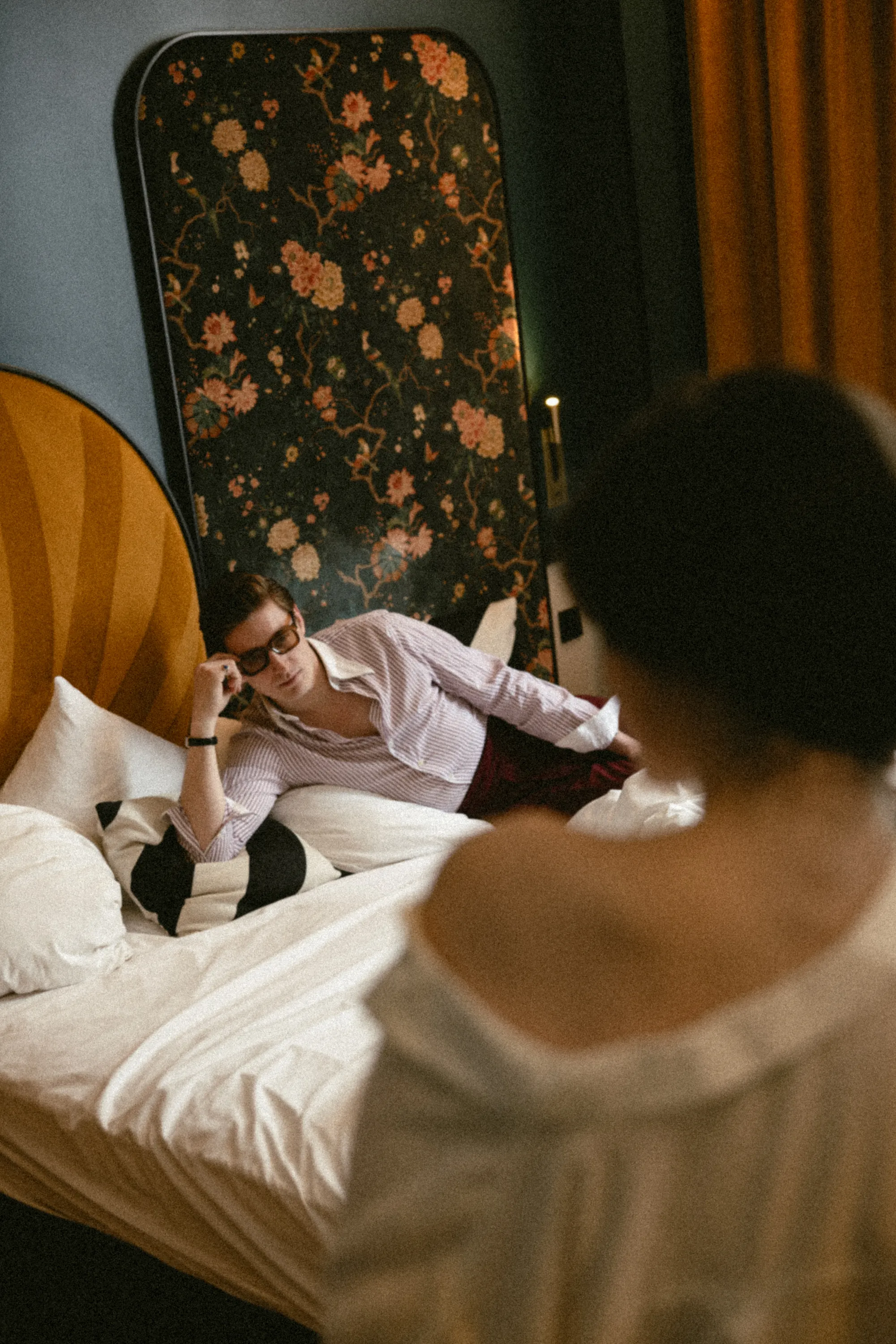 Homme à lunettes allongé sur le lit à tête florale, regardant une personne de dos au premier plan flouté