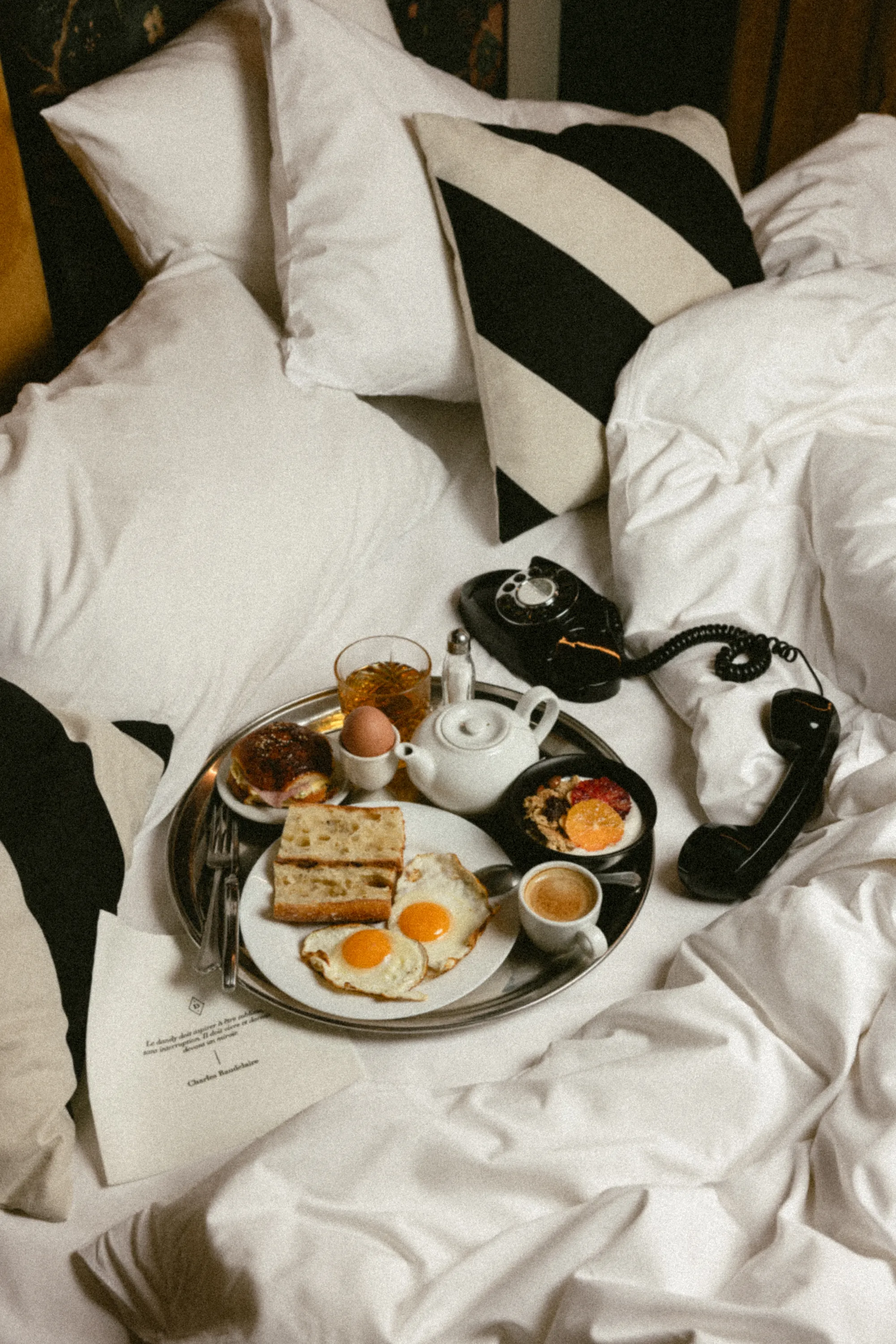 Petit-déjeuner sur plateau argenté posé sur un lit défait, œufs au plat et théière blanche à côté d'un téléphone vintage décroché