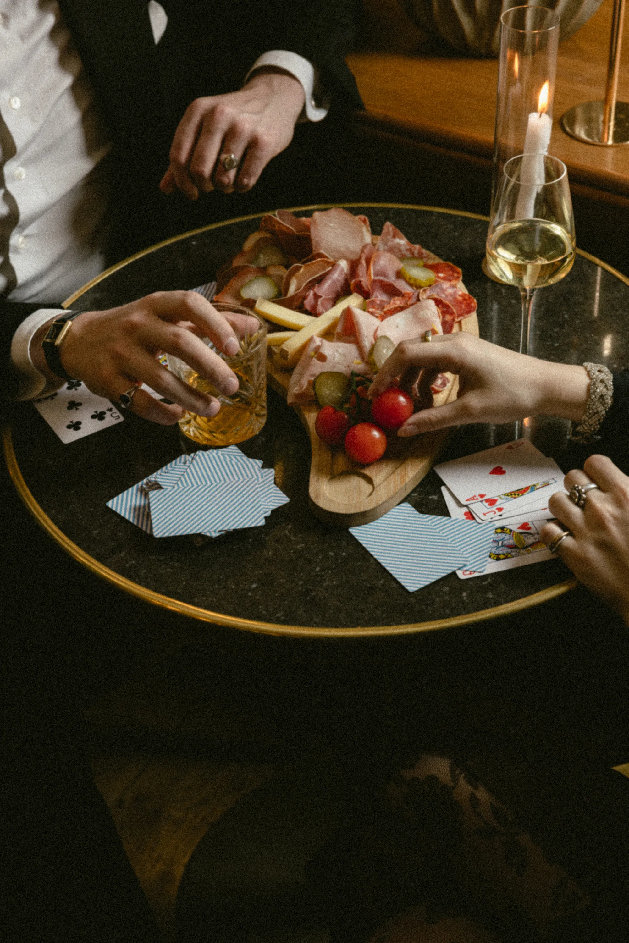 Trois convives partageant une planche de charcuterie et des cartes à jouer autour de verres de vin et de whisky à la bougie