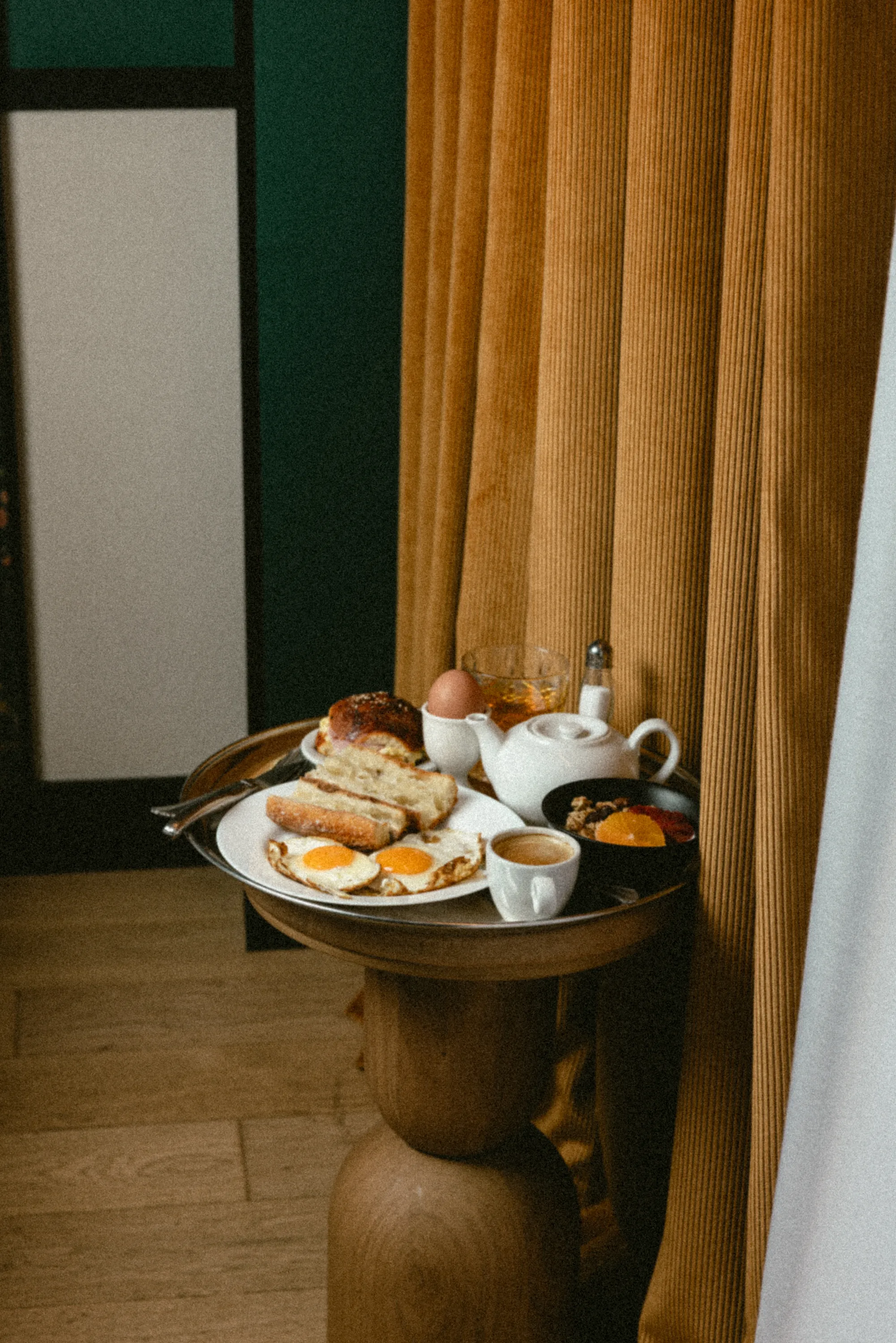 un plateau de petit-déjeuner sur guéridon en bois, œufs et théière blanche devant un rideau moutarde