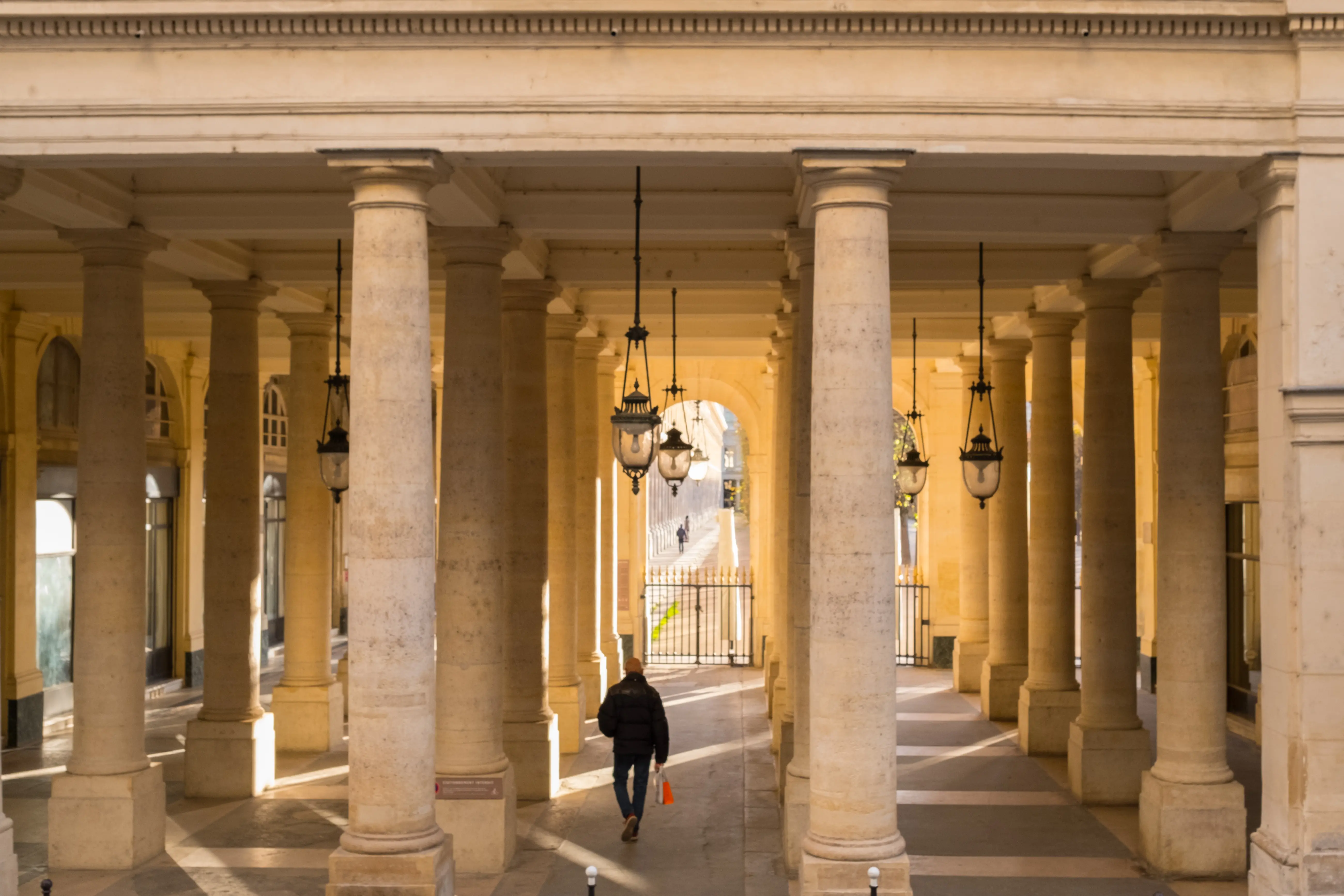 Colonnade dorée baignée de soleil au Palais-Royal, promeneur solitaire marchant sous les lanternes suspendues