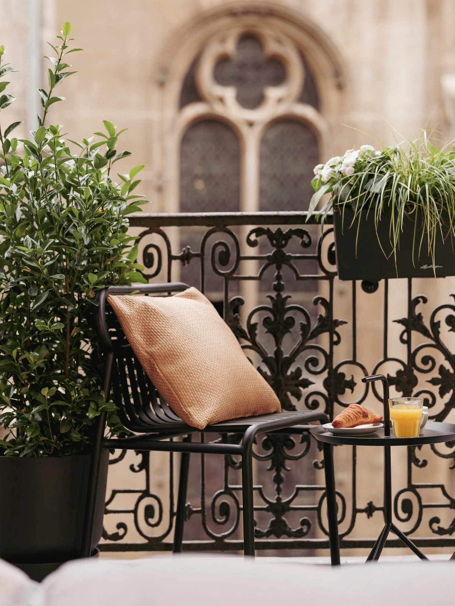 Balcon en fer forgé fleuri de plantes vertes, fauteuil noir à coussin beige et petite table avec croissant et jus d'orange, face à une façade gothique