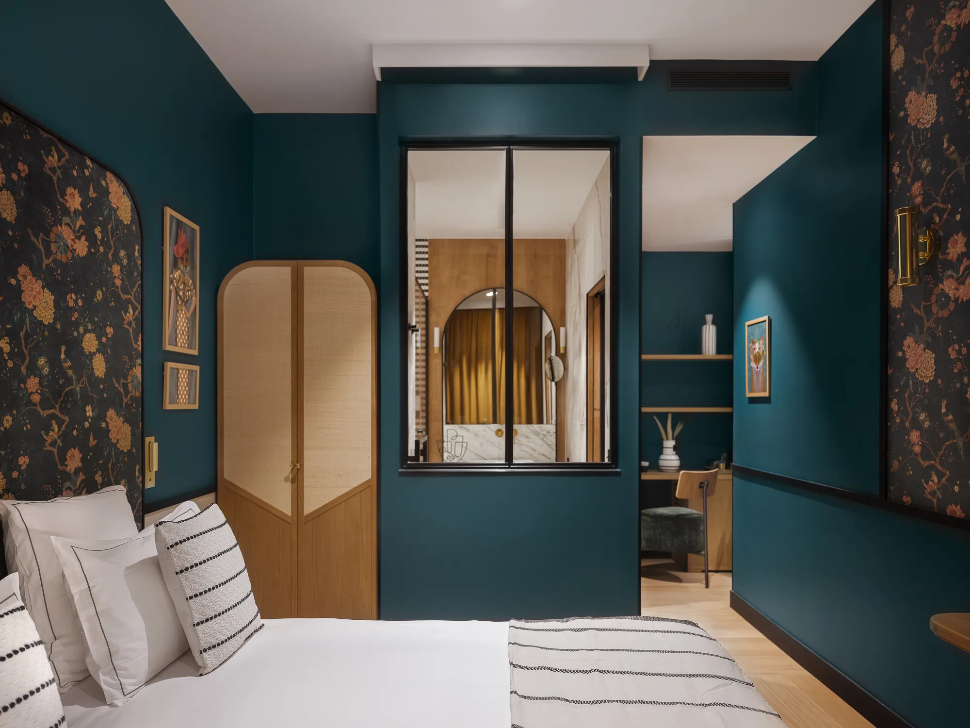 Chambre bleu paon avec verrière intérieure vitrée, penderie en bois et rotin, papier peint floral et coin bureau visible à travers la vitre