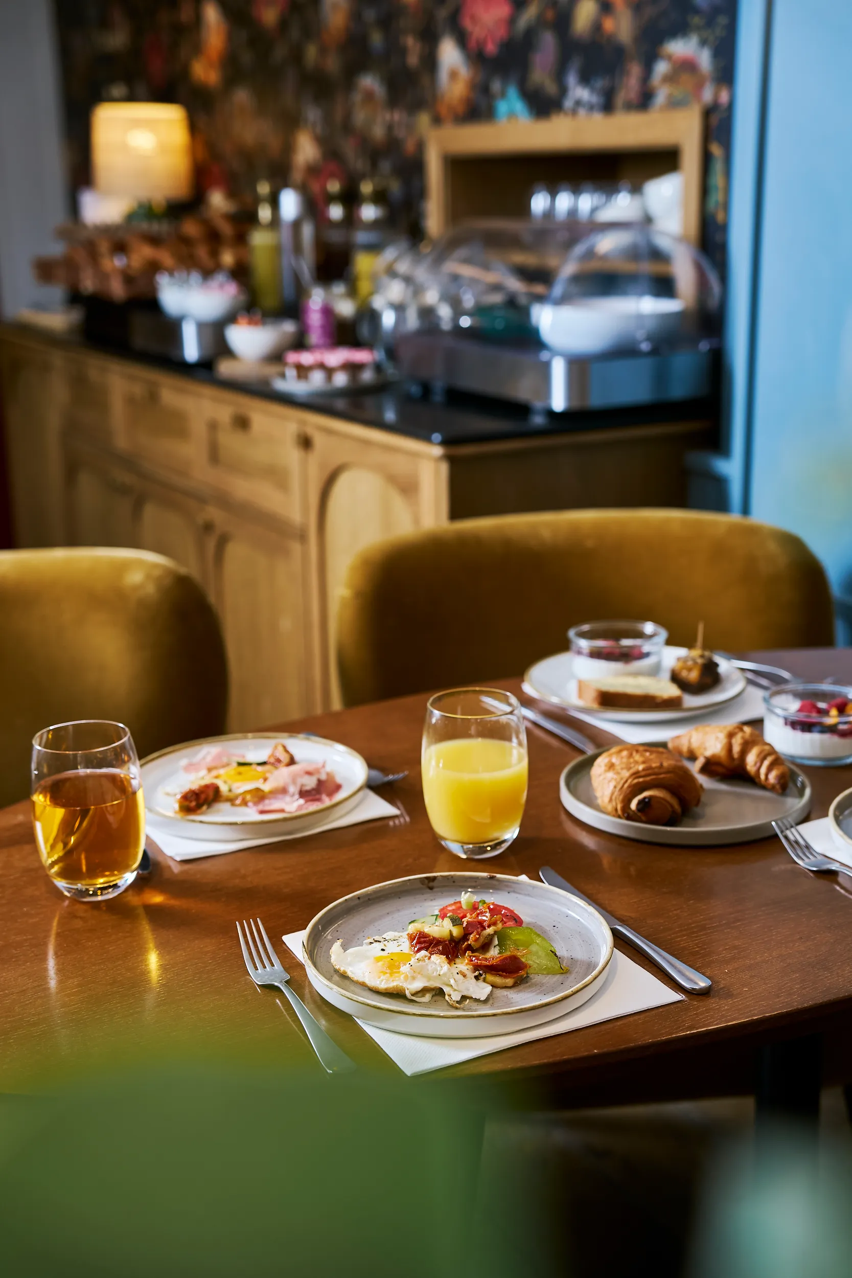 une table de petit-déjeuner en bois avec assiettes garnies et jus d'orange, fauteuils moutarde et buffet en arrière-plan