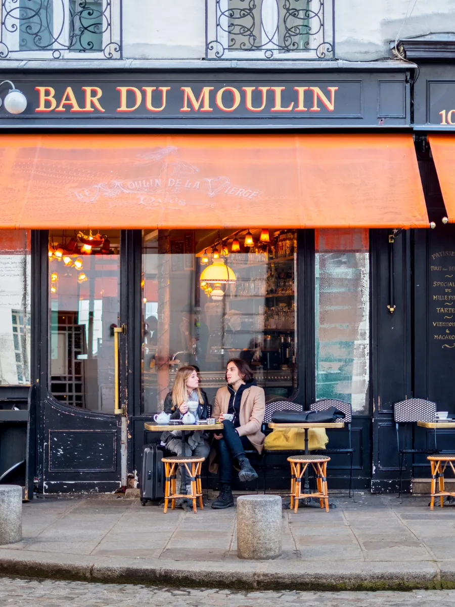 这张巴黎街景照片展现了“Bar du Moulin”咖啡馆阴暗的门面,上方悬挂着一幅鲜艳的橙色遮阳篷。一名年轻男子和一名年轻女子坐在露台的桌边,一边喝着咖啡一边交谈。现场氛围温馨,充满典型的巴黎咖啡馆风情。