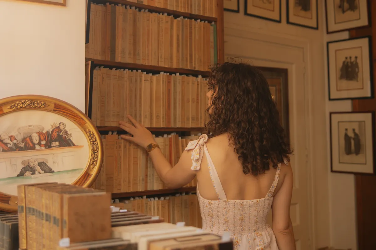 Une femme vue de dos cherchait un livre dans une bibliothèque avec des livres anciens et tableaux aux murs.