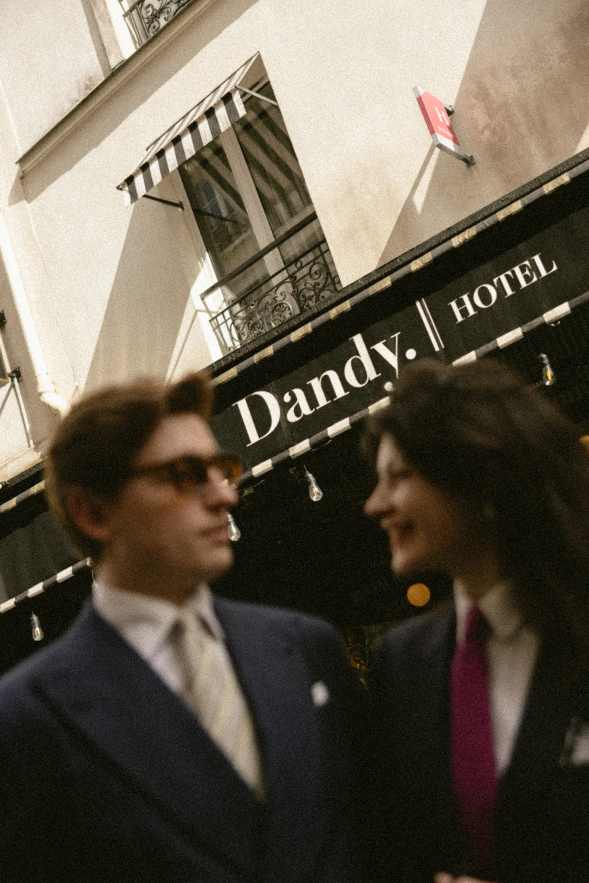 Deux personnes habillées élégamment devant un hôtel nommé Dandy, avec une façade de style parisien et une marquise rayée.