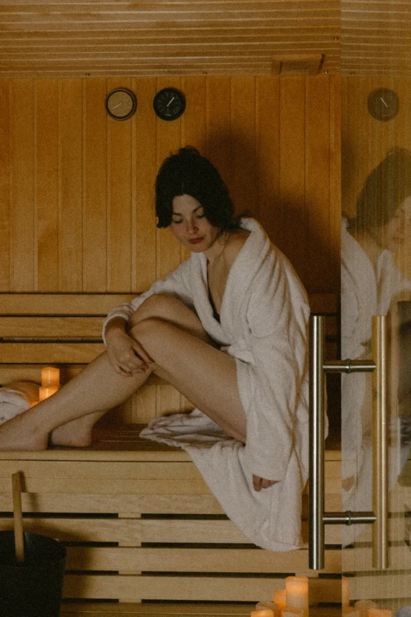 Femme assise dans un sauna en peignoir blanc avec bougies allumées, ambiance relaxante et bois clair sur les murs et bancs.