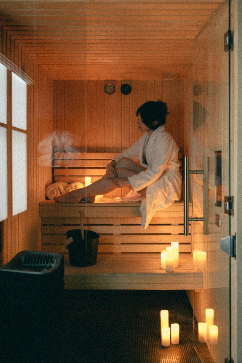 Personne en peignoir blanc assise dans un sauna en bois, entourée de bougies allumées créant une ambiance relaxante.