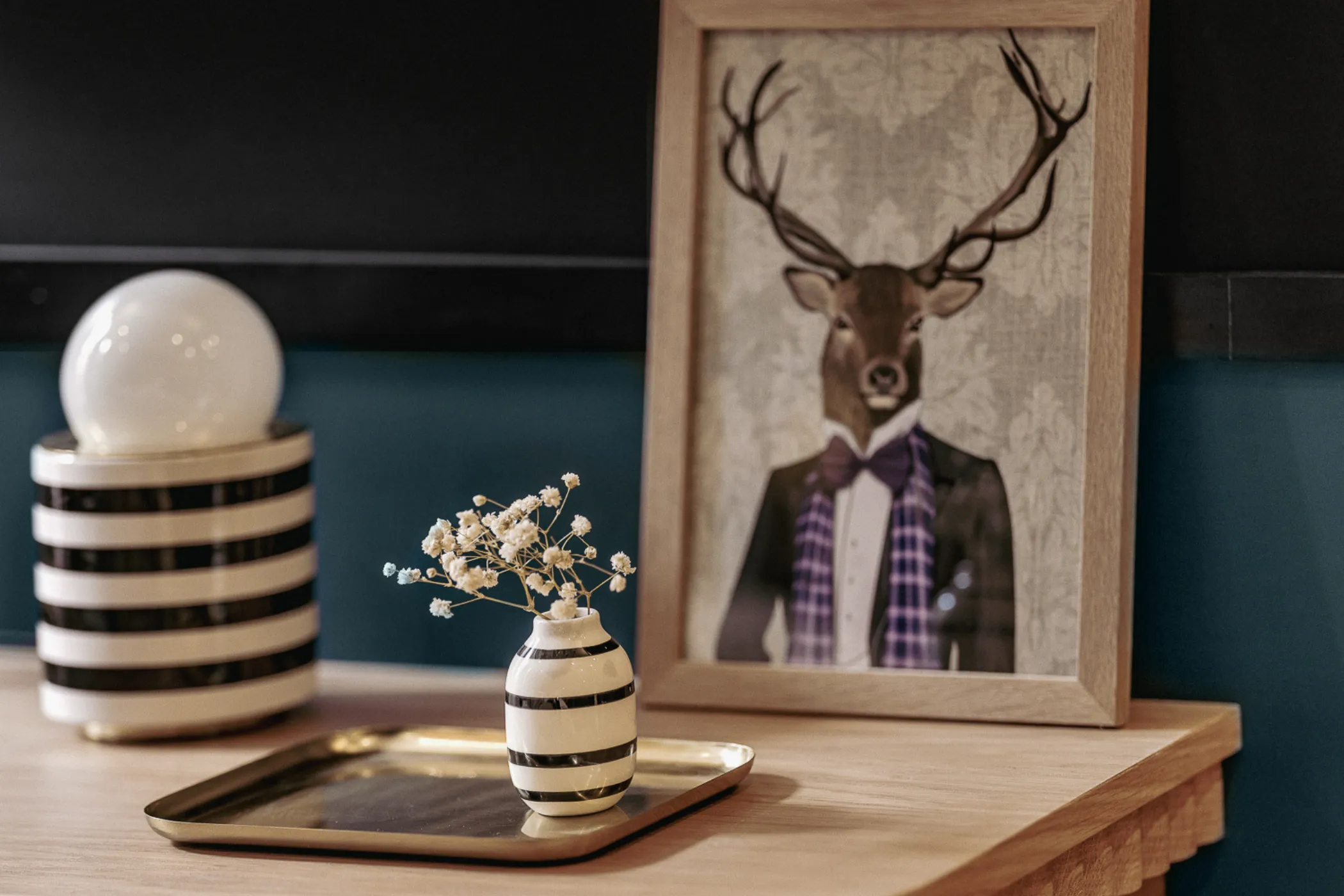 Vase rayé noir et blanc avec de petites fleurs blanches sur un plateau doré posé sur une table en bois, devant un cadre avec une image d'un cerf habillé d'un costume et d'un nœud papillon.