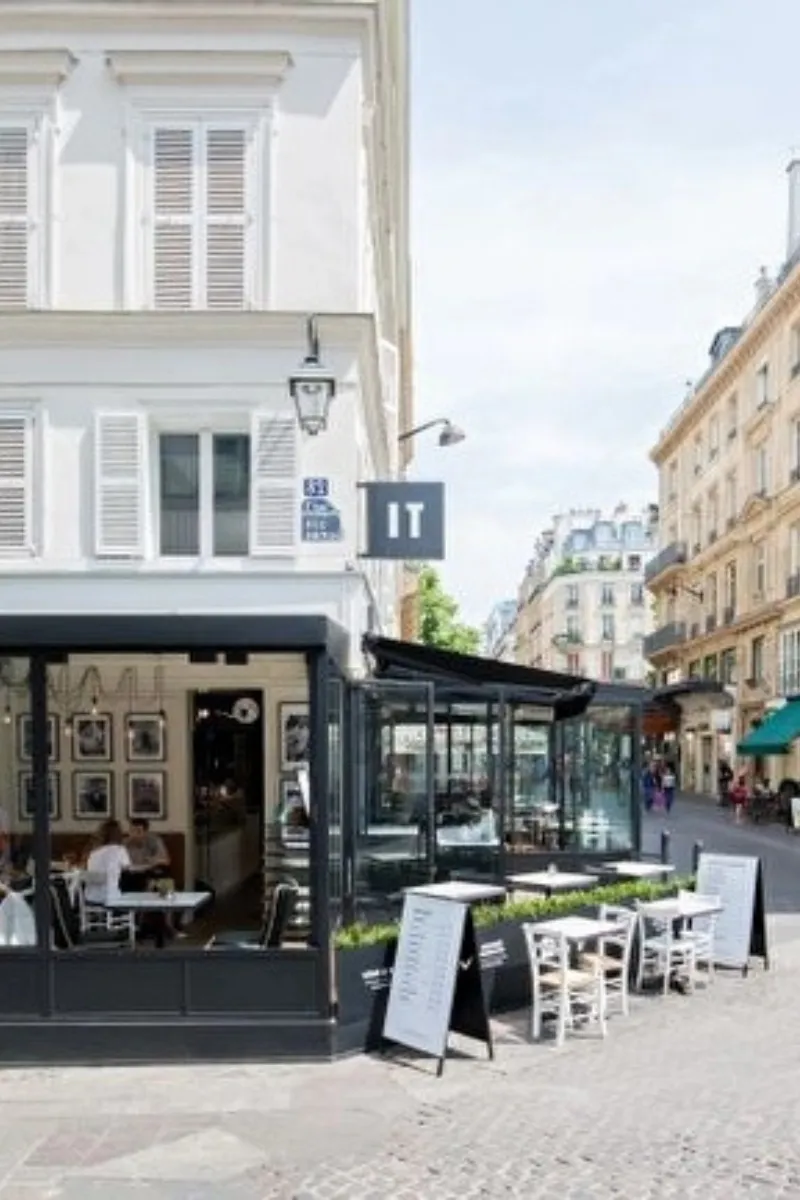 Café avec terrasse extérieure équipée de tables blanches et chaises sur une rue pavée bordée de bâtiments de style parisien, avec panneaux de menu visibles.