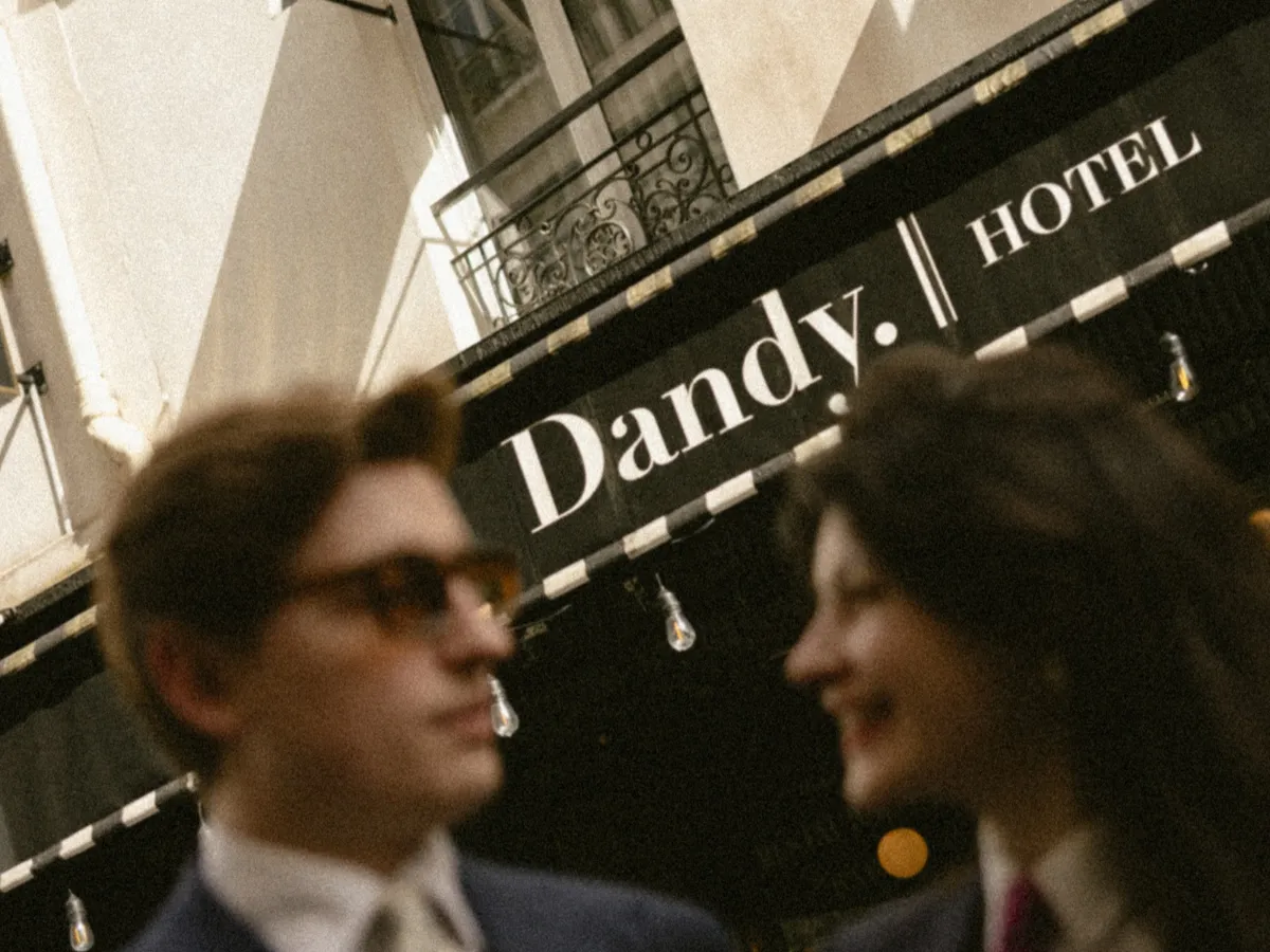 Deux personnes floues en avant-plan discutent devant une enseigne noire avec le texte blanc « Dandy. | HOTEL » sur fond d'architecture urbaine claire.