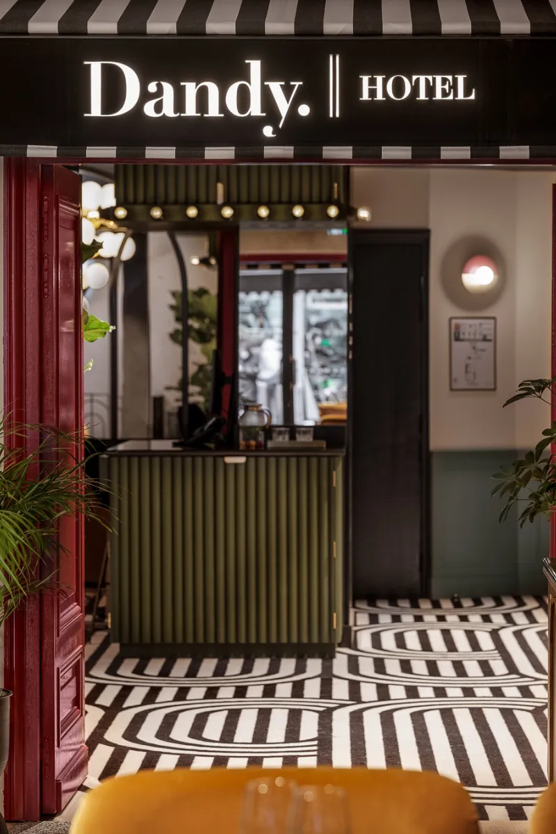 Entrée d'un hôtel avec un panneau lumineux indiquant Dandy. Hotel au-dessus d'une réception verte, sol noir et blanc à motifs géométriques, murs blancs et détails rouges.