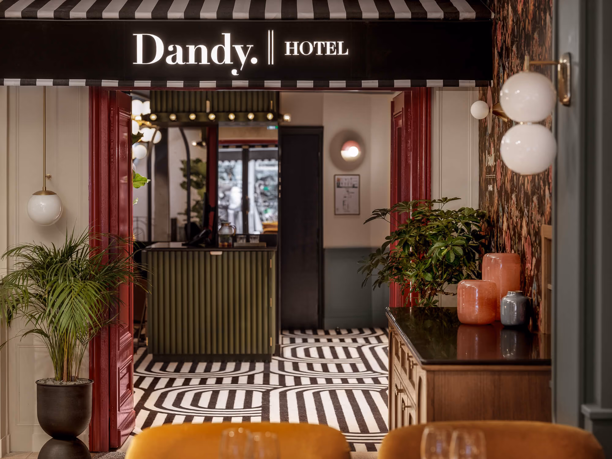 Hall d'entrée de l'hôtel Dandy avec un sol à rayures noires et blanches, un meuble en bois décoré de plantes et de vases, et une réception au fond encadrée par une porte rouge.