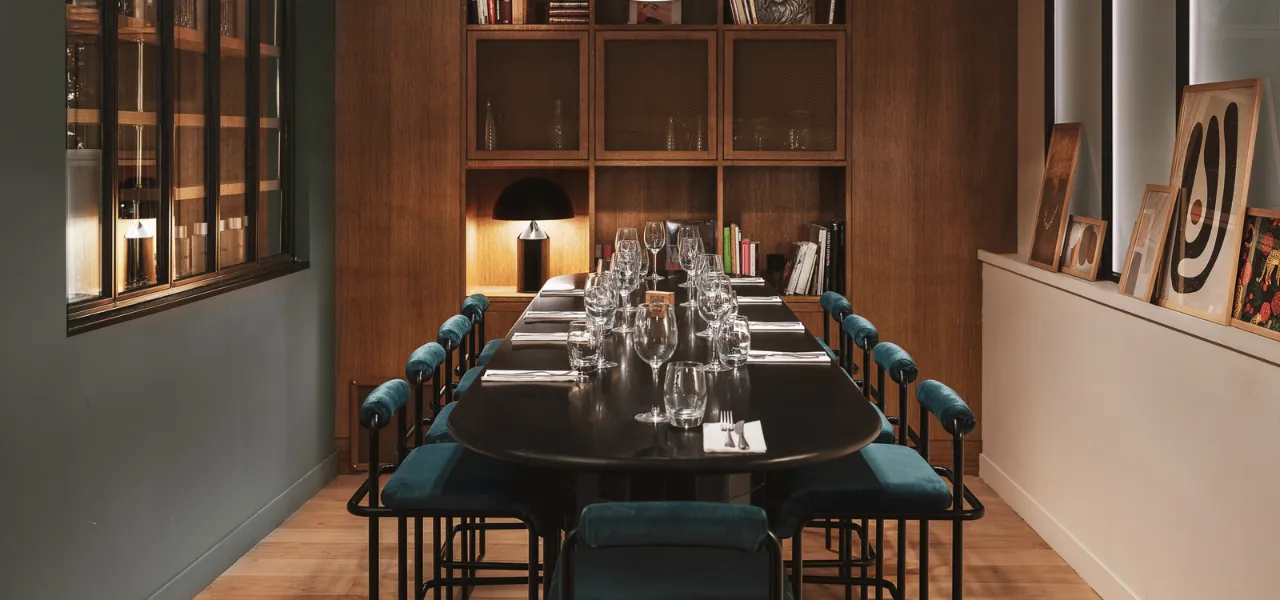 Salle à manger avec une table noire allongée, huit chaises avec coussins bleus, des verres et couverts disposés, une bibliothèque en bois avec lampes et livres, et des cadres décoratifs posés sur un rebord près de la fenêtre.