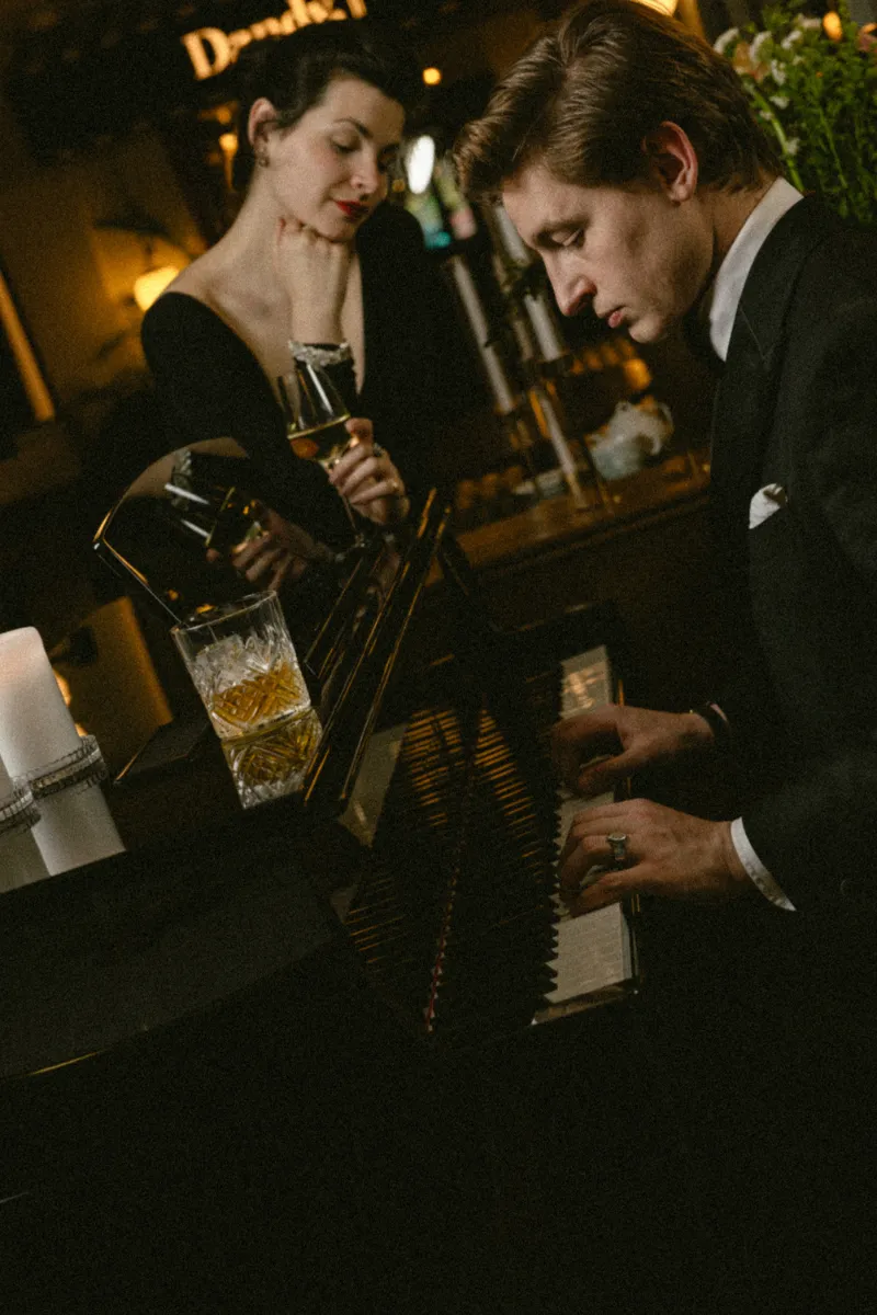 Un homme en costume joue du piano pendant qu'une femme en robe noire l'écoute en tenant un verre de vin dans un cadre chaleureux et intime.