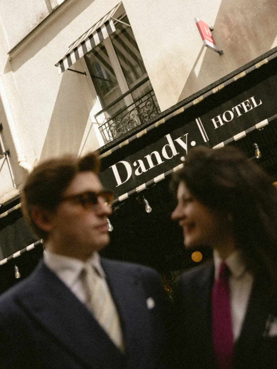 Deux personnes habillées en costume debout et souriant devant un bâtiment avec une enseigne indiquant « Dandy. HOTEL ».
