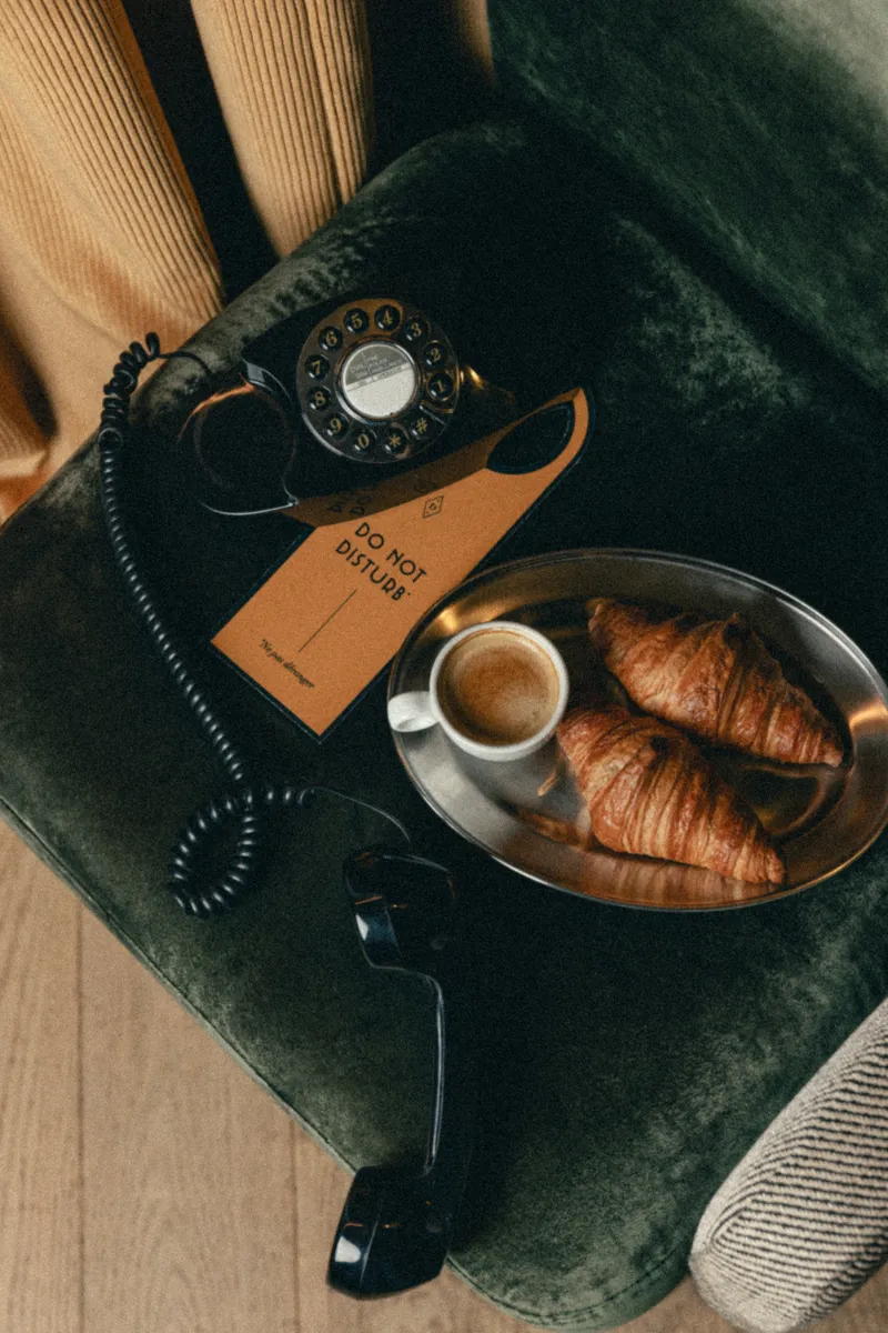Téléphone noir ancien à cadran rotatif avec le combiné décroché sur un fauteuil vert, une pancarte « Ne pas déranger », et un plateau avec un café espresso et deux croissants.